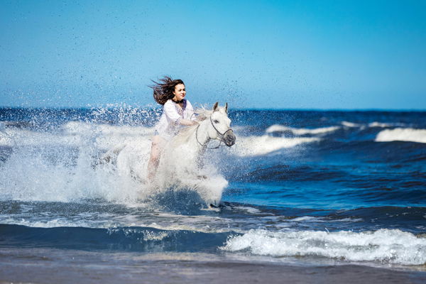 Girl on a white horse storming through the water on the seashore. Galopade. Horseback riding. (photocreo)