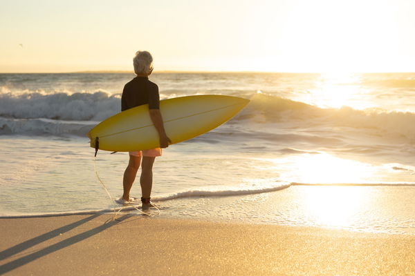 Old woman with a surfboard at the beach Rear view of a senior Caucasian woman at the beach at sunset